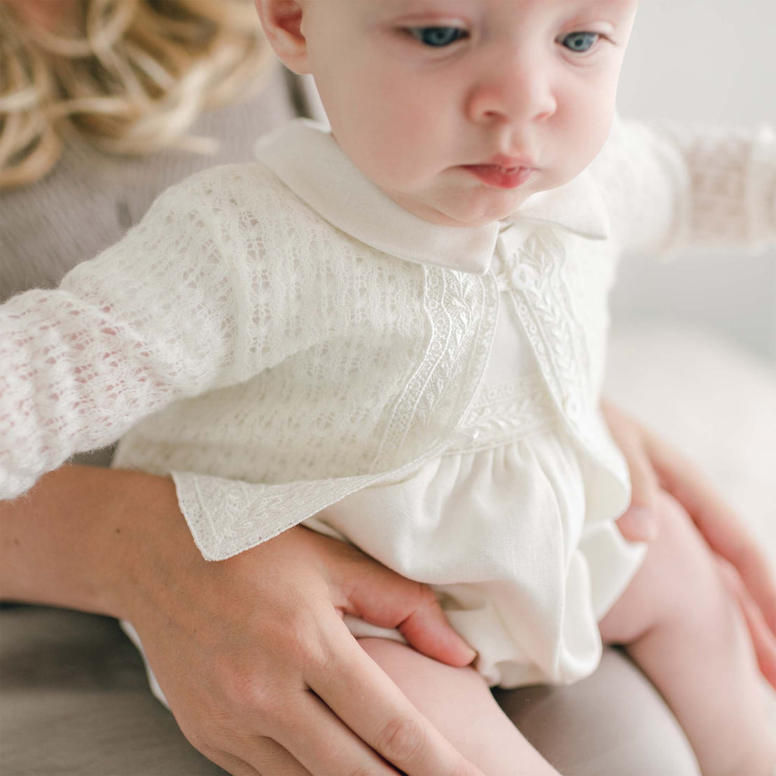 A close-up of a baby dressed in an Oliver Knit Sweater with lacy detailing and a collar, sitting on an adult's lap. The baby boy's comfort is evident, with the adult's hands gently supporting him from behind. The background is softly blurred, focusing on the baby's calm expression.