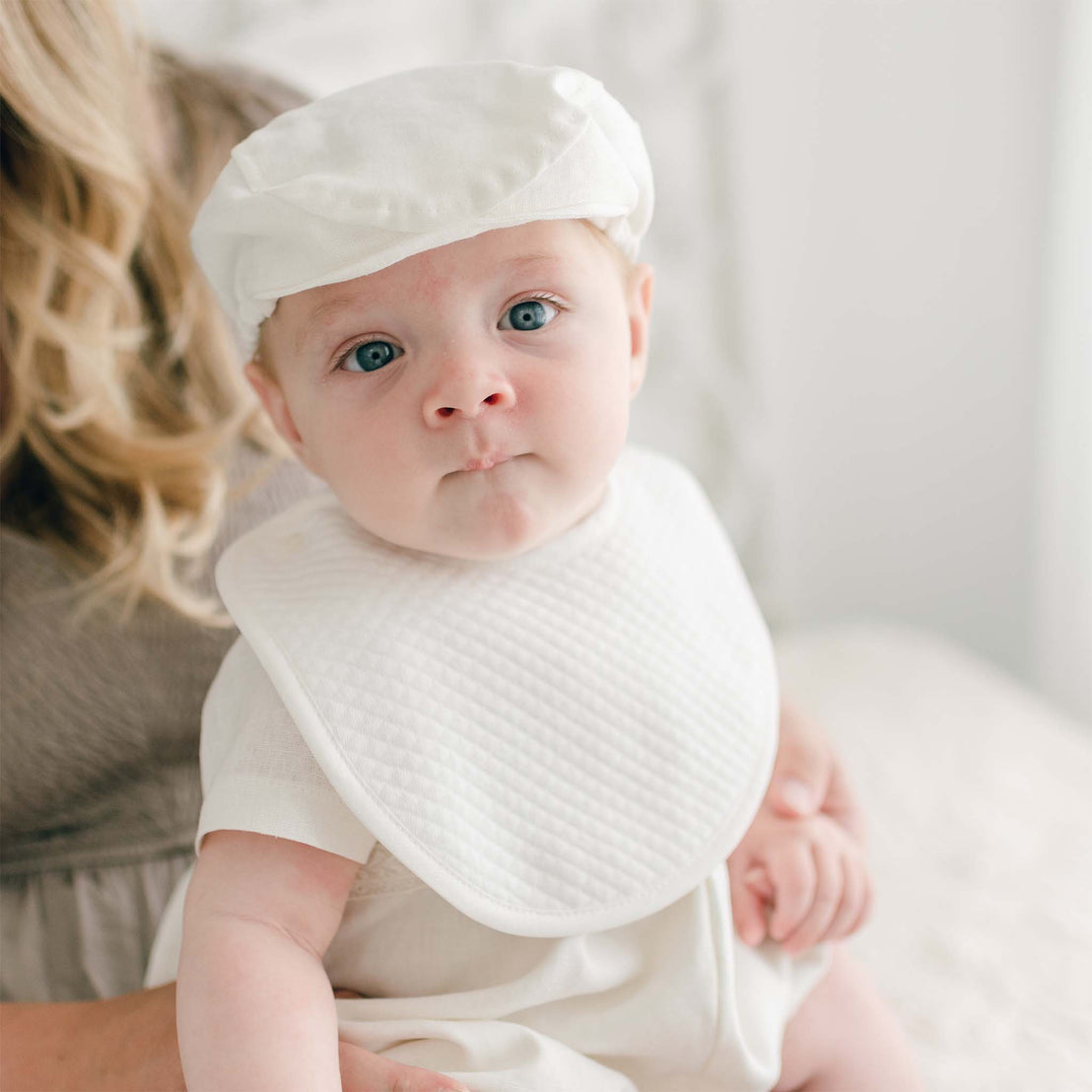 A baby with blue eyes wearing a white cap and an Oliver Bib is sitting on a woman's lap. The woman's face is not visible, and she has blonde, wavy hair. The baby looks directly at the camera with a slightly pouting expression.