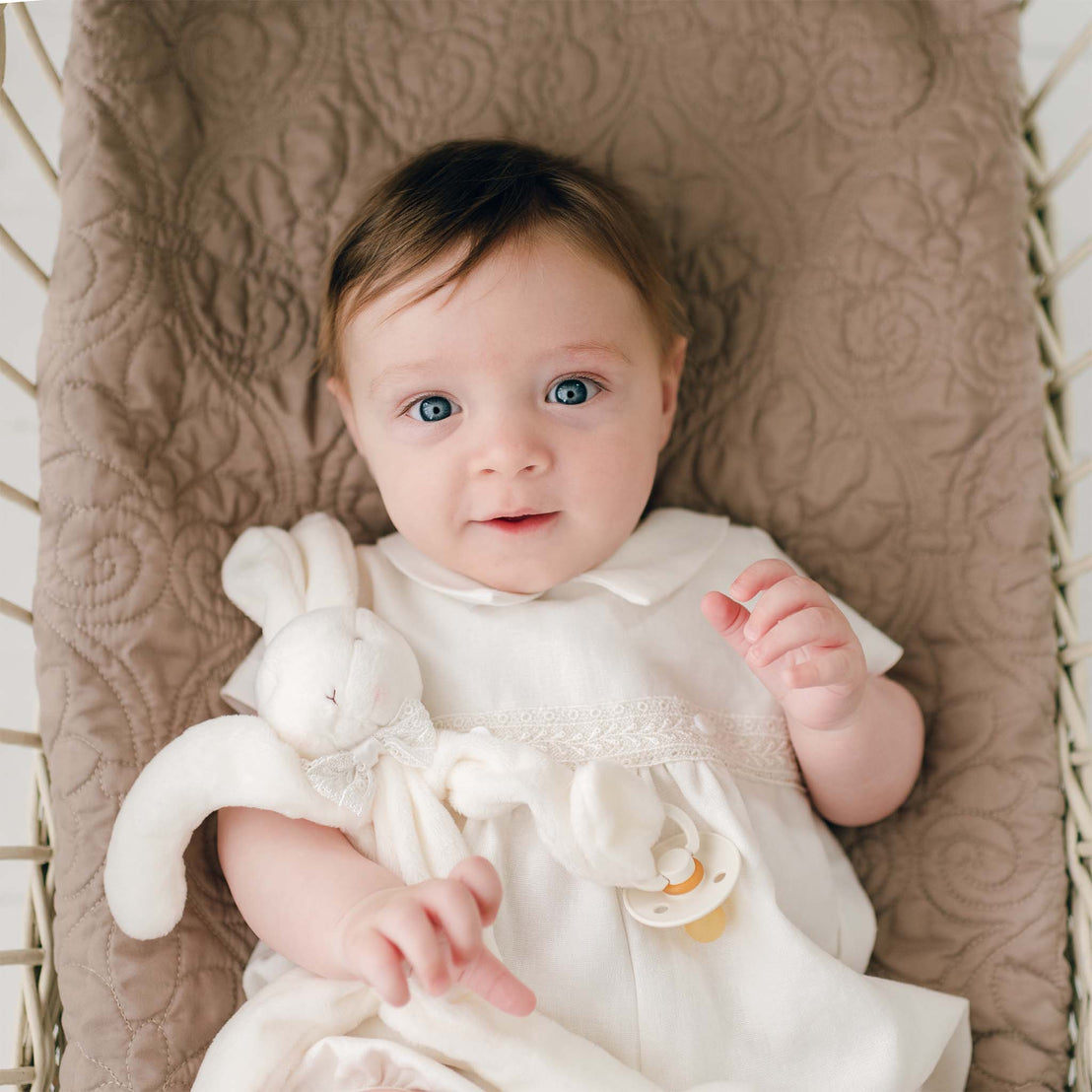 A baby boy wearing the Oliver Romper in ivory. He is holding a Oliver Silly Bunny Buddy in his right arm. His eyes show a curious expression as he gazes up.