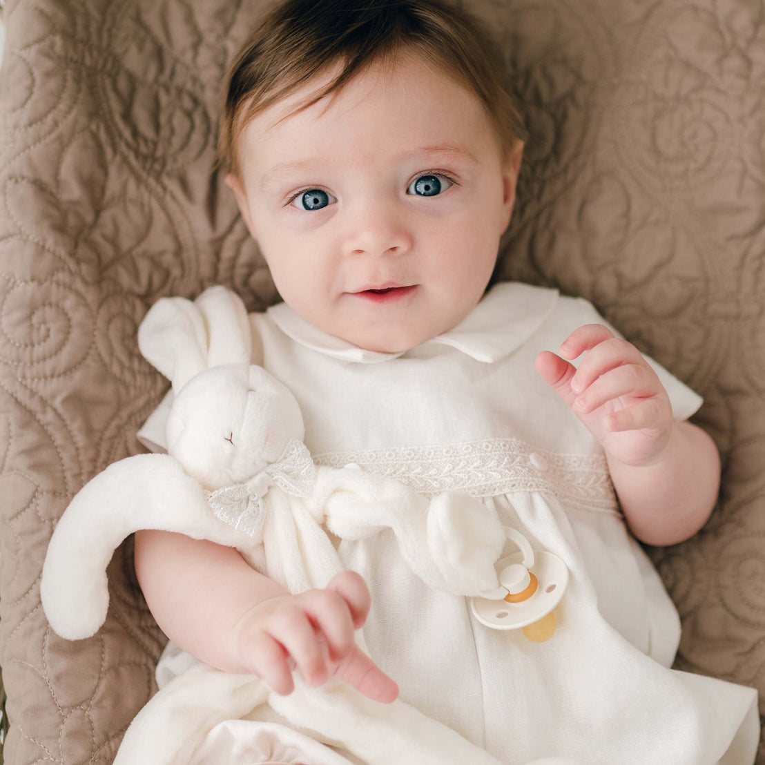 A baby with light brown hair and blue eyes lies on a quilted beige blanket, wearing the Oliver Romper in white. The baby holds an Oliver Silly Bunny Buddy | Pacifier Holder made of soft white velour and has a baby-safe pacifier clip attached to its hand. The baby has a calm, curious expression.