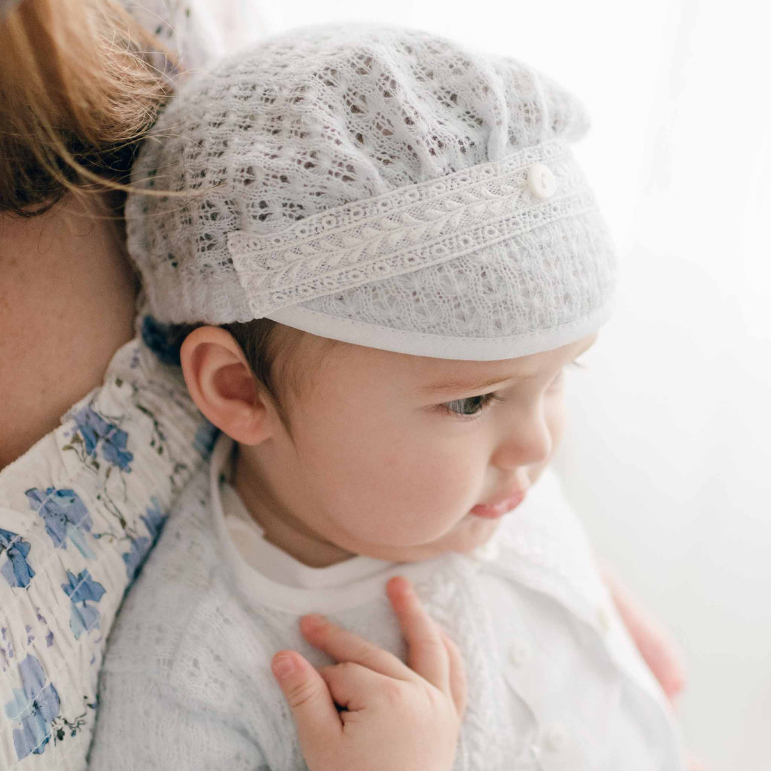 A baby dressed in the Oliver Romper and Knit Sweater and Cap ensemble in white featuring intricate embroidery. He is cradled by a partially visible adult, while he gazes to the side as light streams through a window in the background.