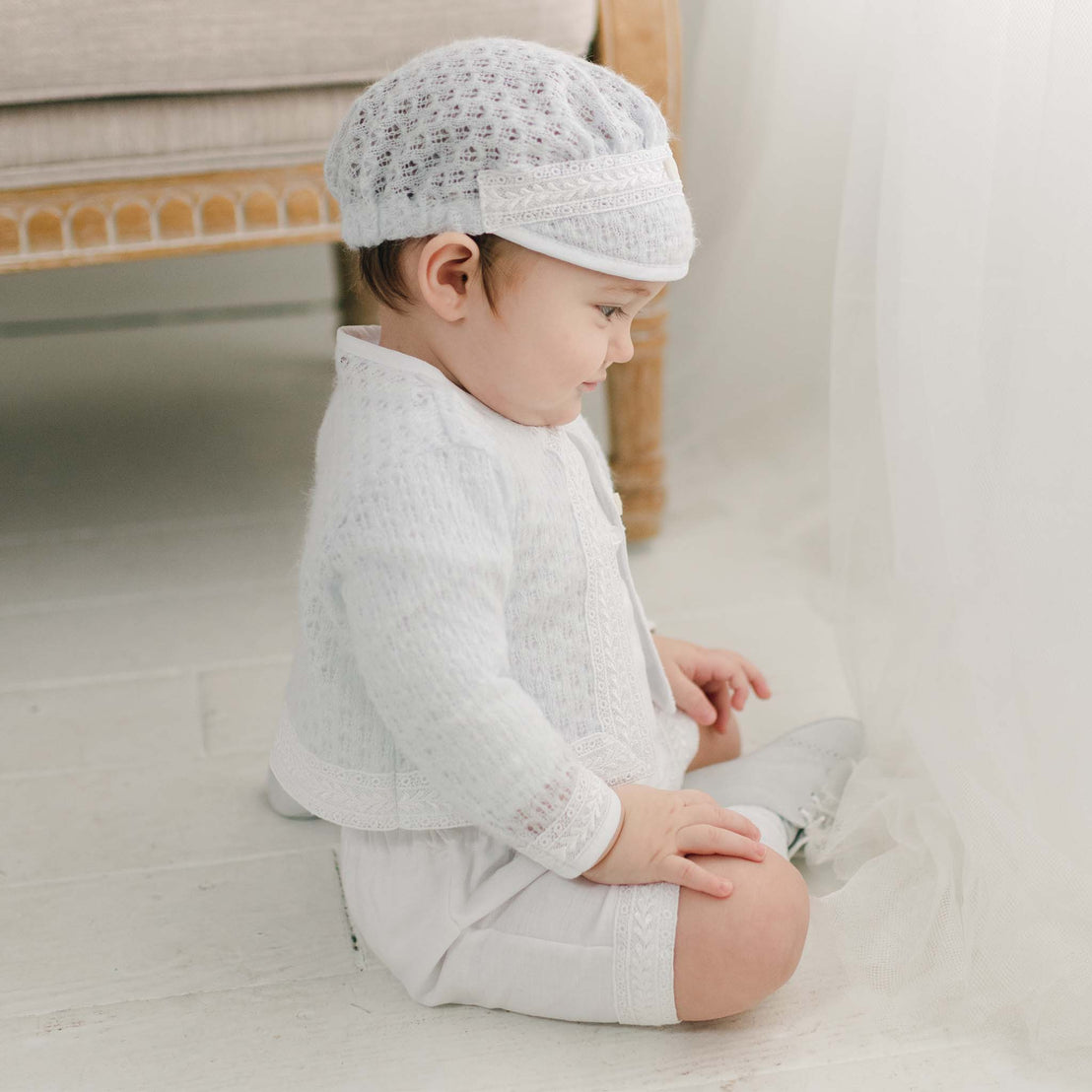 A baby dressed in the Oliver Romper and Knit Sweater and Cap ensemble in white featuring intricate embroidery. He is sitting on the floor and looking down at something near their hands. In the background, there is a beige chair and a sheer white curtain.