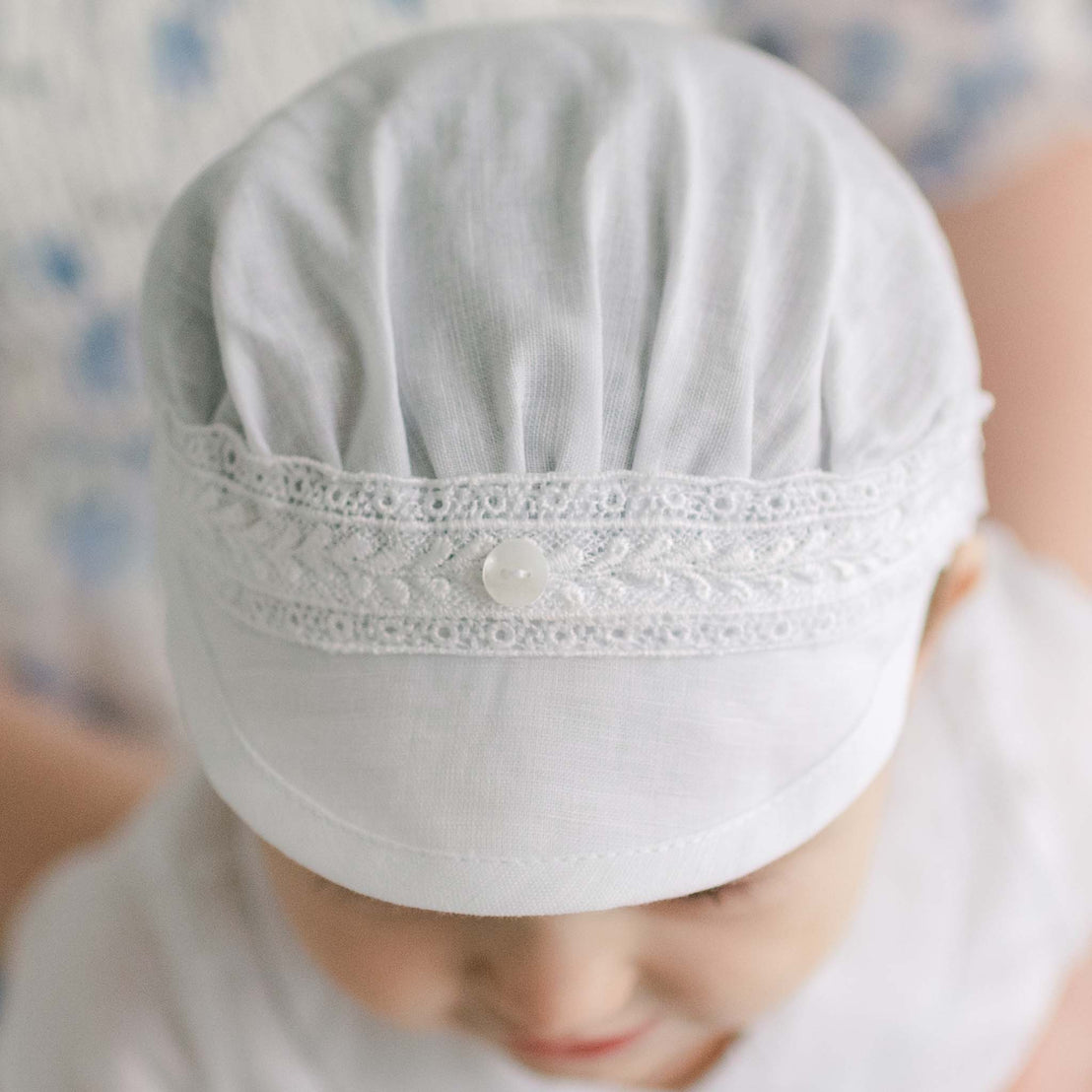 Baby boy wearing a white Oliver Linen Cap. The hat is made from linen and features a Venice lace across the front and a button for added detail.