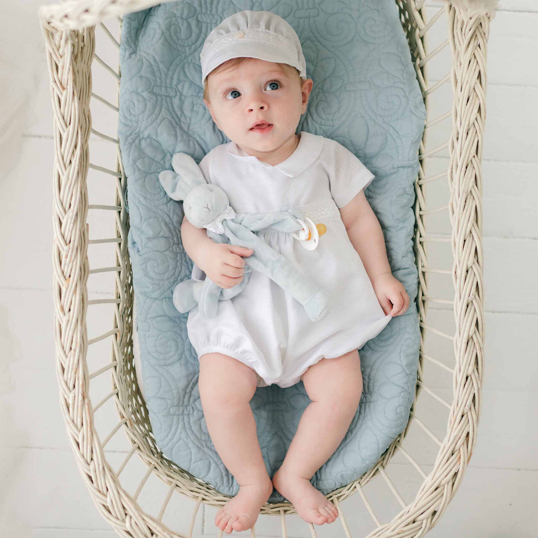 A baby boy wearing the Oliver Romper and matching linen cap in white. He clutches the Oliver Bunny Buddy pacifier holder in his right arm. The boy is laying in a wicker basket that is lined with a blue fleur-de-lys quilted blanket. The baby boy gazes up with a curious glance.