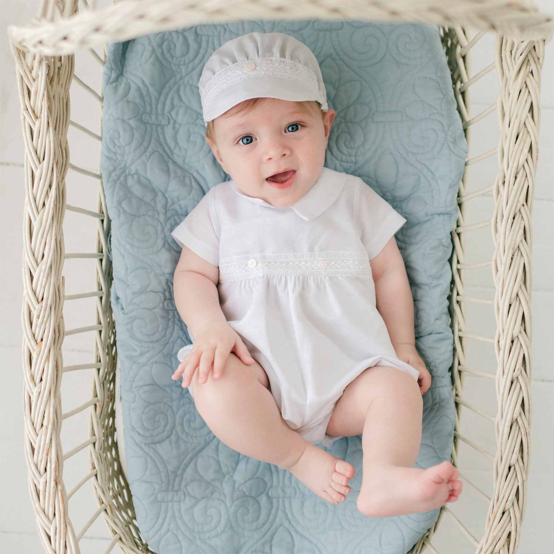 A baby boy wearing the Oliver Romper in white with matching linen cap. He is laying in a wicker basket lined with a blue fleur-de-lys quilted blanket. He appears to be looking up with a happy expression.