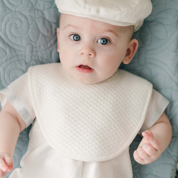 A baby boy wearing the Owen Bib over the Owen Linen Romper lies on a teal quilted cotton blanket. The baby's blue eyes look up with a curious expression.