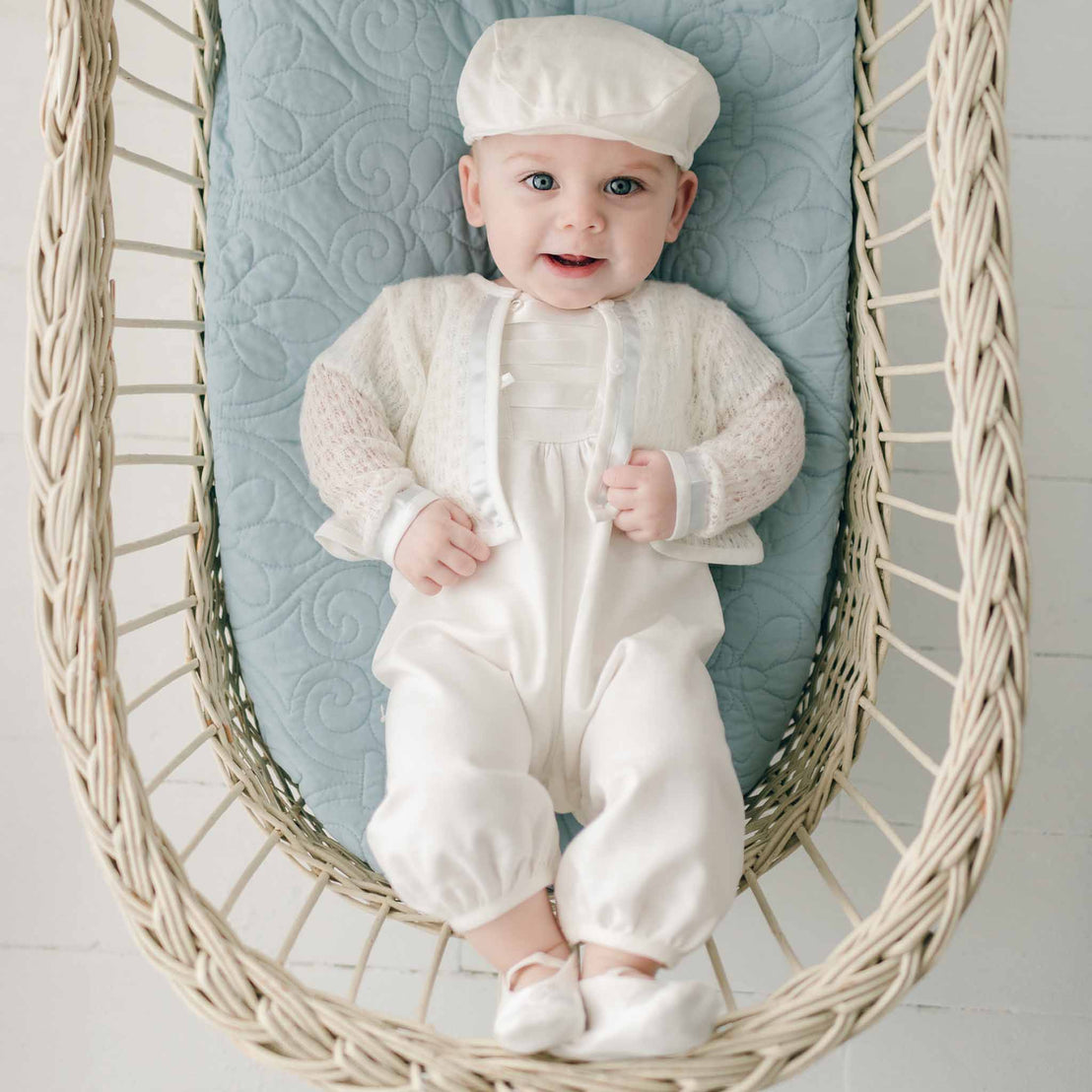 A cheerful baby dressed in an Owen Knit Sweater and cap, adorned with a blue silk ribbon, lying in a woven hanging basket, looking up with a bright smile.