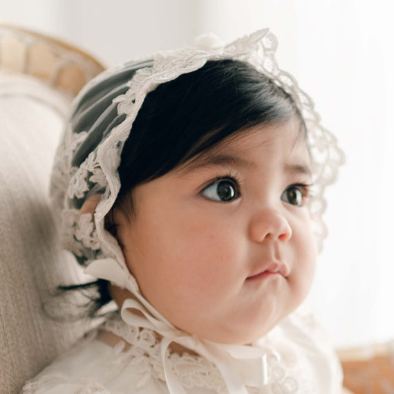 Baby girl wearing the Penelope Lace Bonnet. The bonnet features an embroidered netting with a floral pattern in ivory and champagne. Her gaze is focused to the side of the camera.