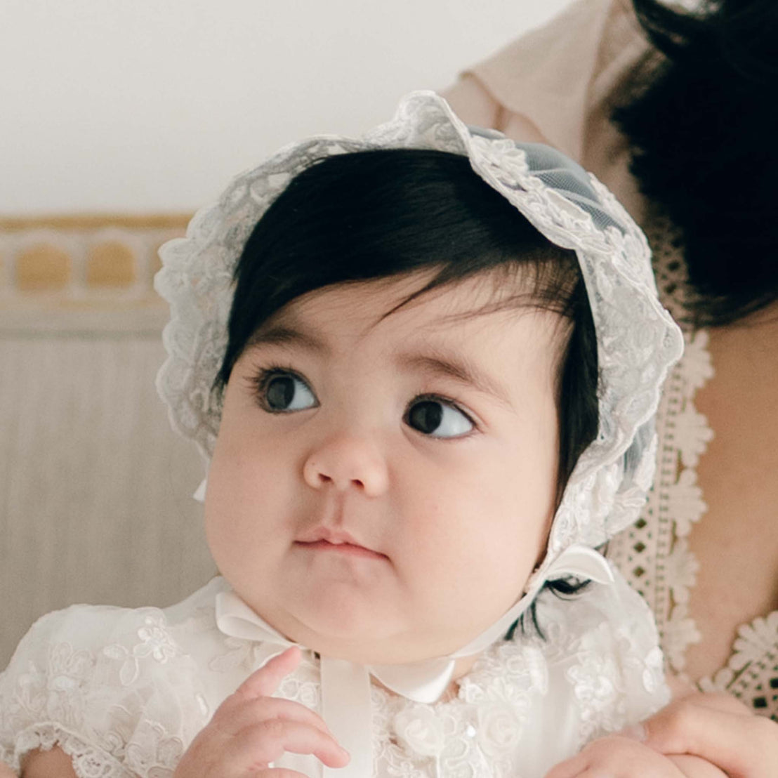 Baby girl with her mother by her side. She is wearing the Penelope Lace Bonnet and matching Penelope Christening Gown. The bonnet features embroidered netting with a floral pattern in ivory and champagne. The background is softly blurred.