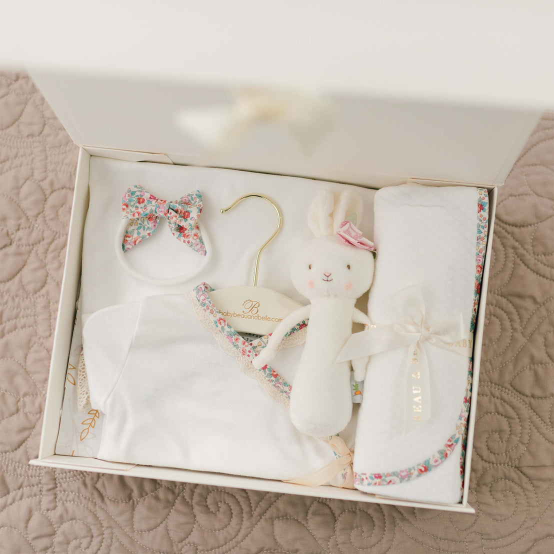 A Petite Fleur Gift Set - Save 10% for a baby, featuring a white outfit with floral trim from Petite Fleur clothing, a matching floral bow hair clip, a personalized baby blanket with similar floral detail, and a white stuffed bunny with floral ears. All items are neatly arranged on a quilted surface.