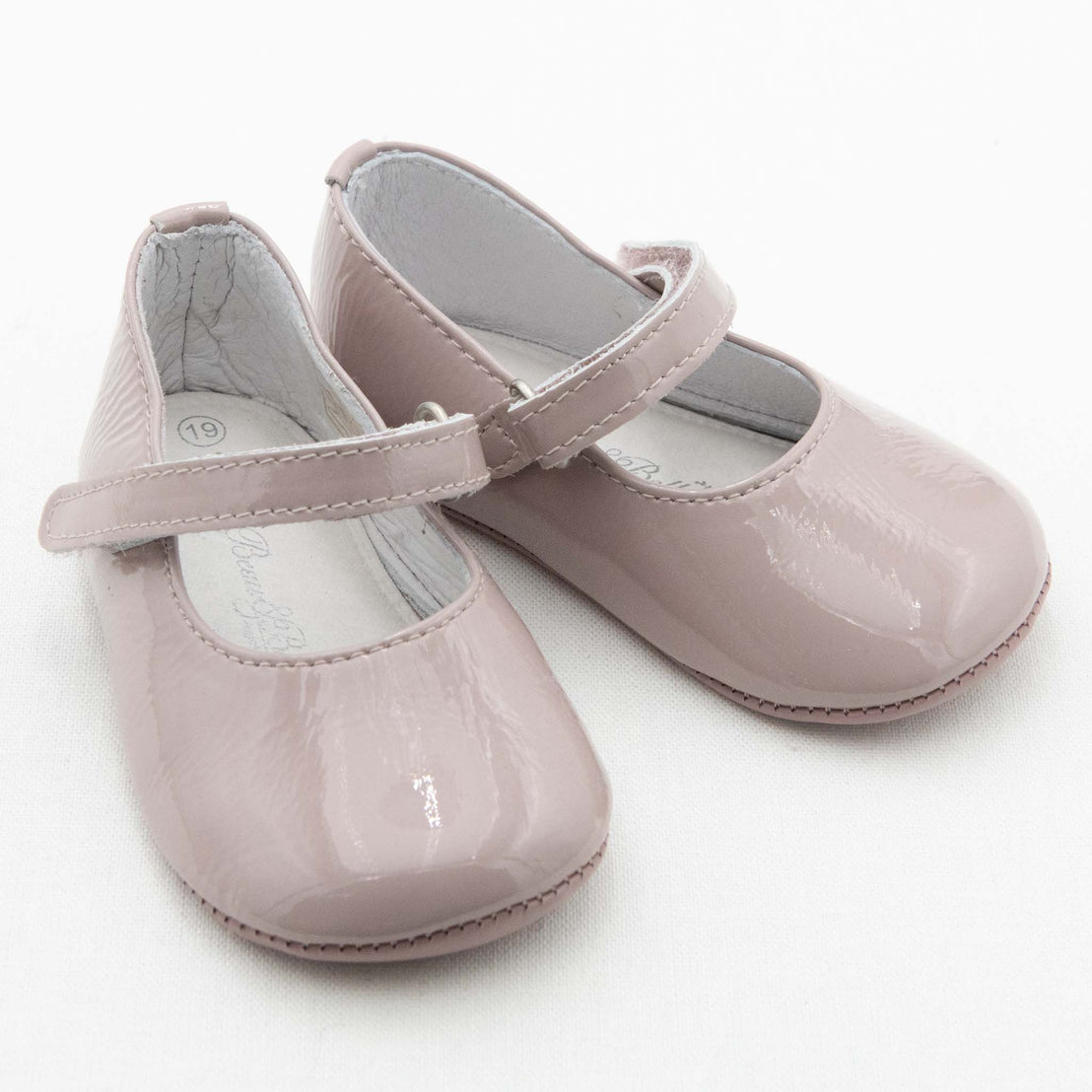 A pair of Rose Patent Leather Mary Janes. The shoes are made with pink patent leather with a soft velcro strap. The coordinating rose pink chiffon bow and feather are removed from the strap. The background is white.