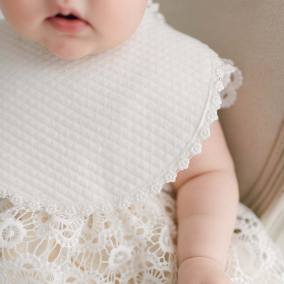 A close-up of a baby wearing a Poppy Bib with a textured pattern and lace edges. The baby's baptism outfit underneath also has lace detailing, adding a vintage touch. The image is focused on the baby's torso, with the face partially visible.