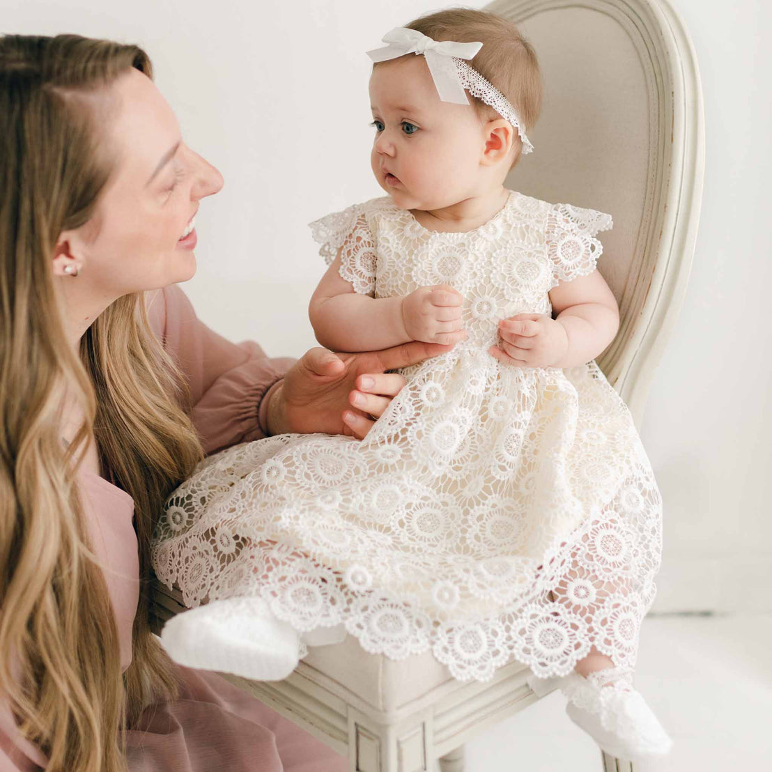 A woman with long blonde hair sits next to a baby on a white chair. The baby, dressed in a Poppy Dress & Bonnet, touches the woman's arm. The woman, dressed in a pink blouse, looks at the baby affectionately.