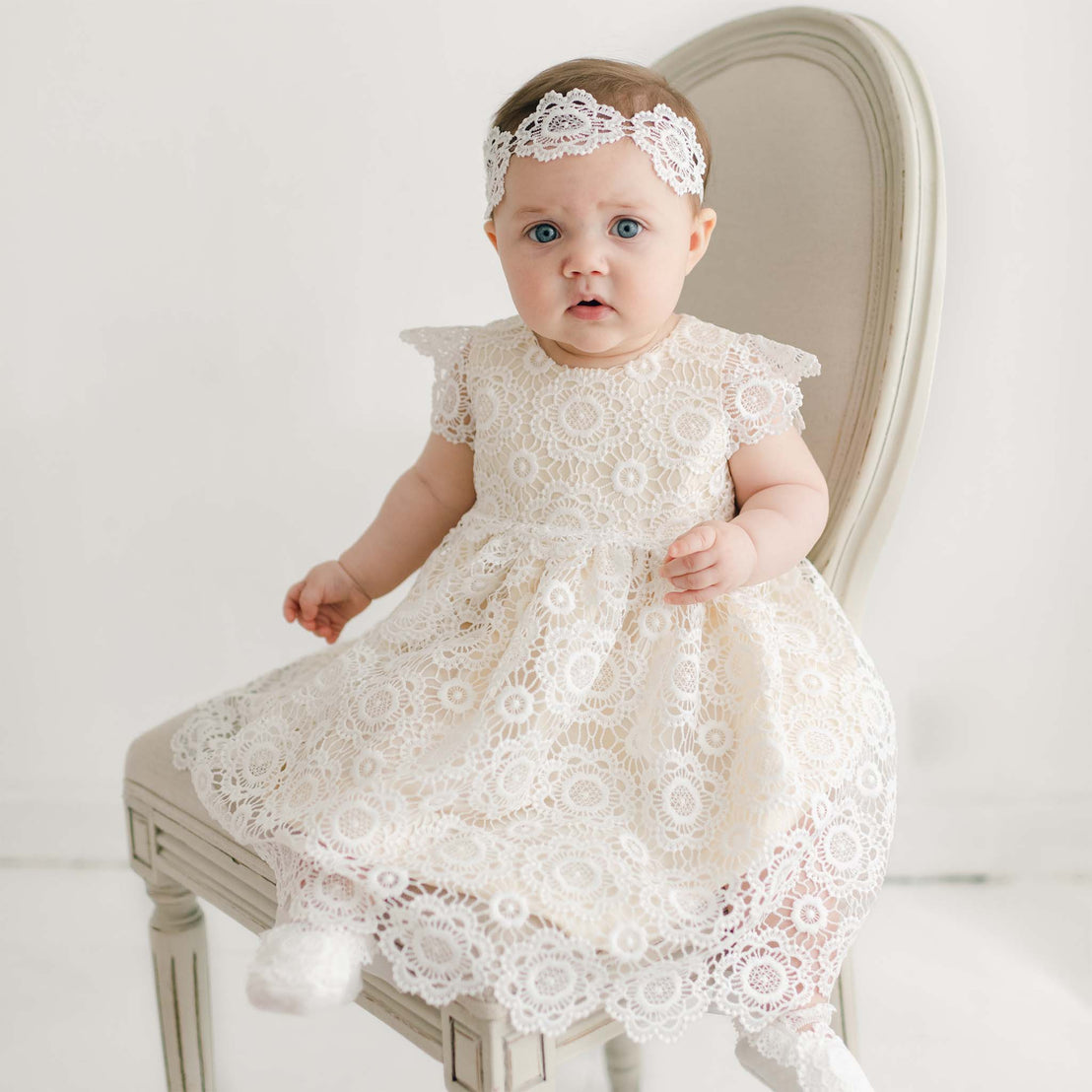 A baby sits on a chair wearing the Poppy Dress & Bonnet. The baby has blue eyes and light brown hair. The background is a simple, light-colored setting, highlighting the exquisitely handmade in the USA attire.