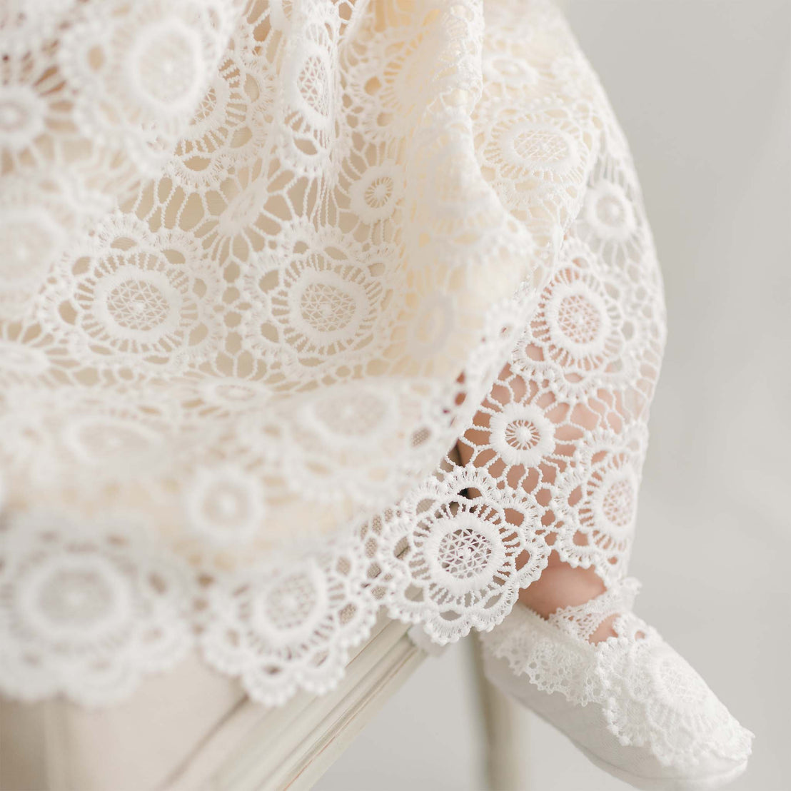 A close-up of a baby dressed in the Poppy Dress & Bonnet, featuring a cream-colored, lace-patterned dress with intricate floral designs and a silk dupioni lining. The baby is seated, with one foot visible, wearing a matching lace shoe. Handmade in the USA, the soft, delicate textures and light tones give the image an airy, elegant feel.