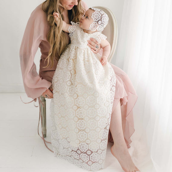 A woman in a flowy pink dress is sitting on a chair, holding a baby dressed in a long, white Poppy Christening Gown & Bonnet. They are both smiling and looking at each other. The background is bright and minimalistic, with sheer white curtains adding a soft touch.