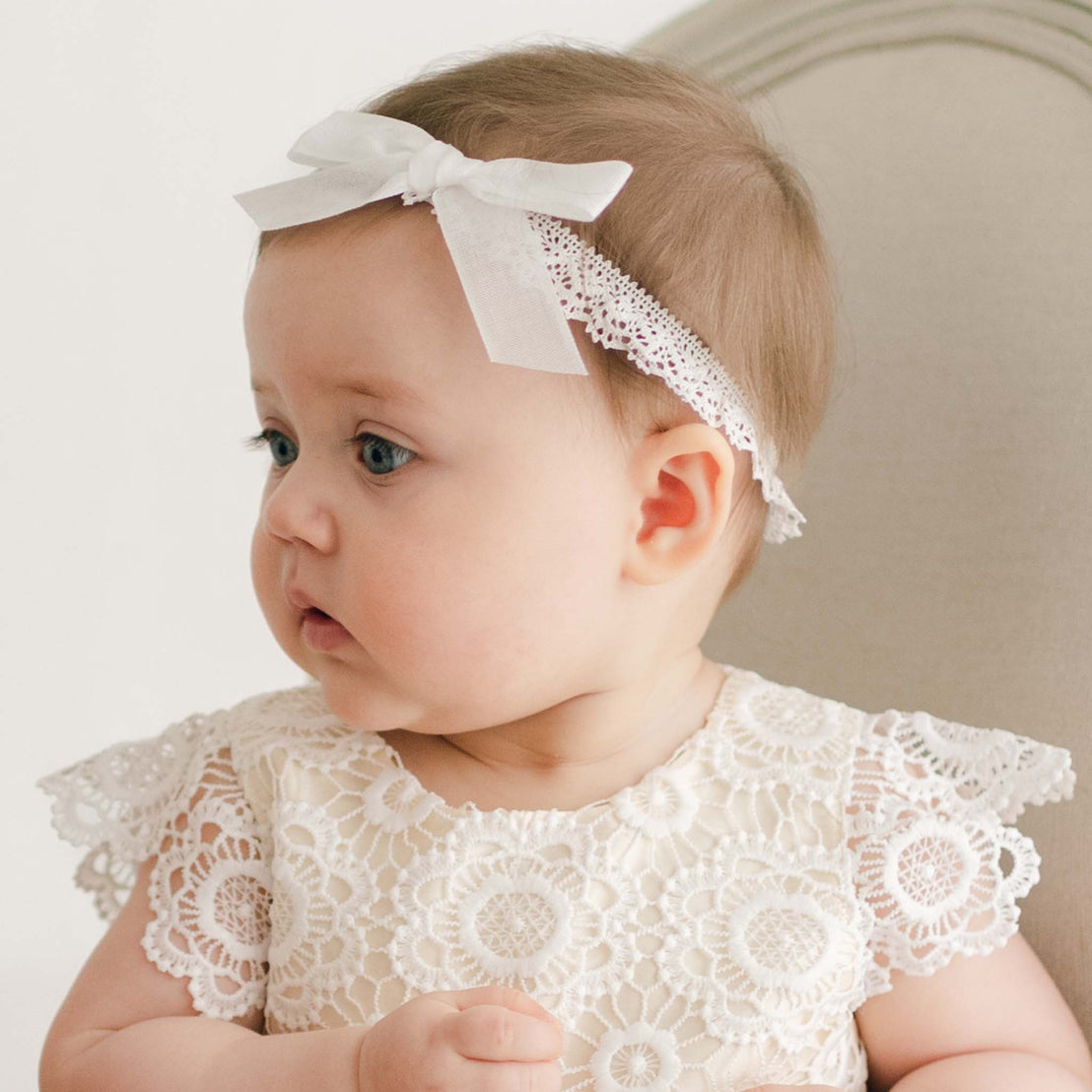 A baby with light brown hair is wearing a Poppy Headband and a delicate white lace top, reminiscent of a christening gown. The baby is looking to the side while sitting in a beige chair. The background is light and minimalistic.