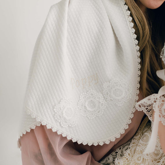 Close-up of a person partially draped in a Poppy Personalized Blanket with intricate lace trim and floral patterns. The name "Poppy" is delicately embroidered in gold on the blanket, making it a perfect personalized gift. The person is wearing a sheer, light pink outfit.