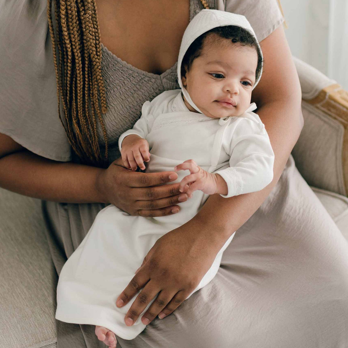 Baby girl sitting with her mother and wearing the Ivory Newborn Quinn Layette Gown and Ivory Quinn Quilted Cotton Bonnet.