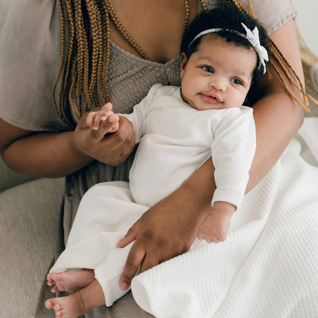 Closer detail of a baby girl sitting with her mother and wearing the Ivory Newborn Quinn Layette Gown and Quinn Lace Bow Headband.