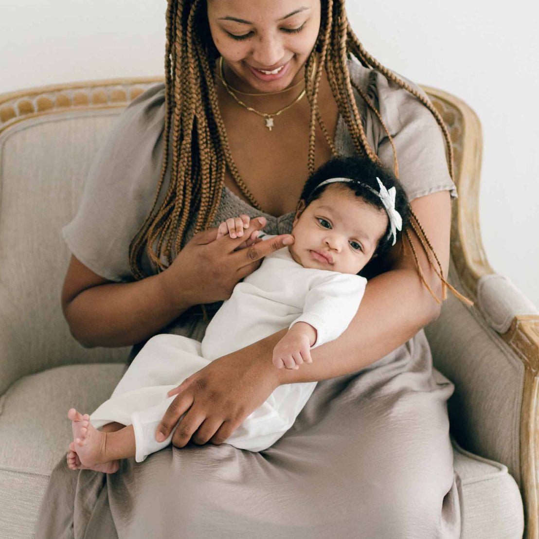 Baby girl sitting with her mother and wearing the Ivory Newborn Quinn Layette Gown and Quinn Lace Bow Headband.