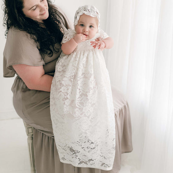 A woman holding a baby dressed in the Rose Christening Gown & Bonnet and looking towards the camera in a softly lit room with sheer curtains.