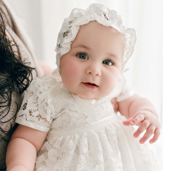 A close-up of a baby with bright blue eyes dressed in the Rose Lace Bonnet and matching Rose Christening Gown, looking directly at the camera with an expression of curiosity.