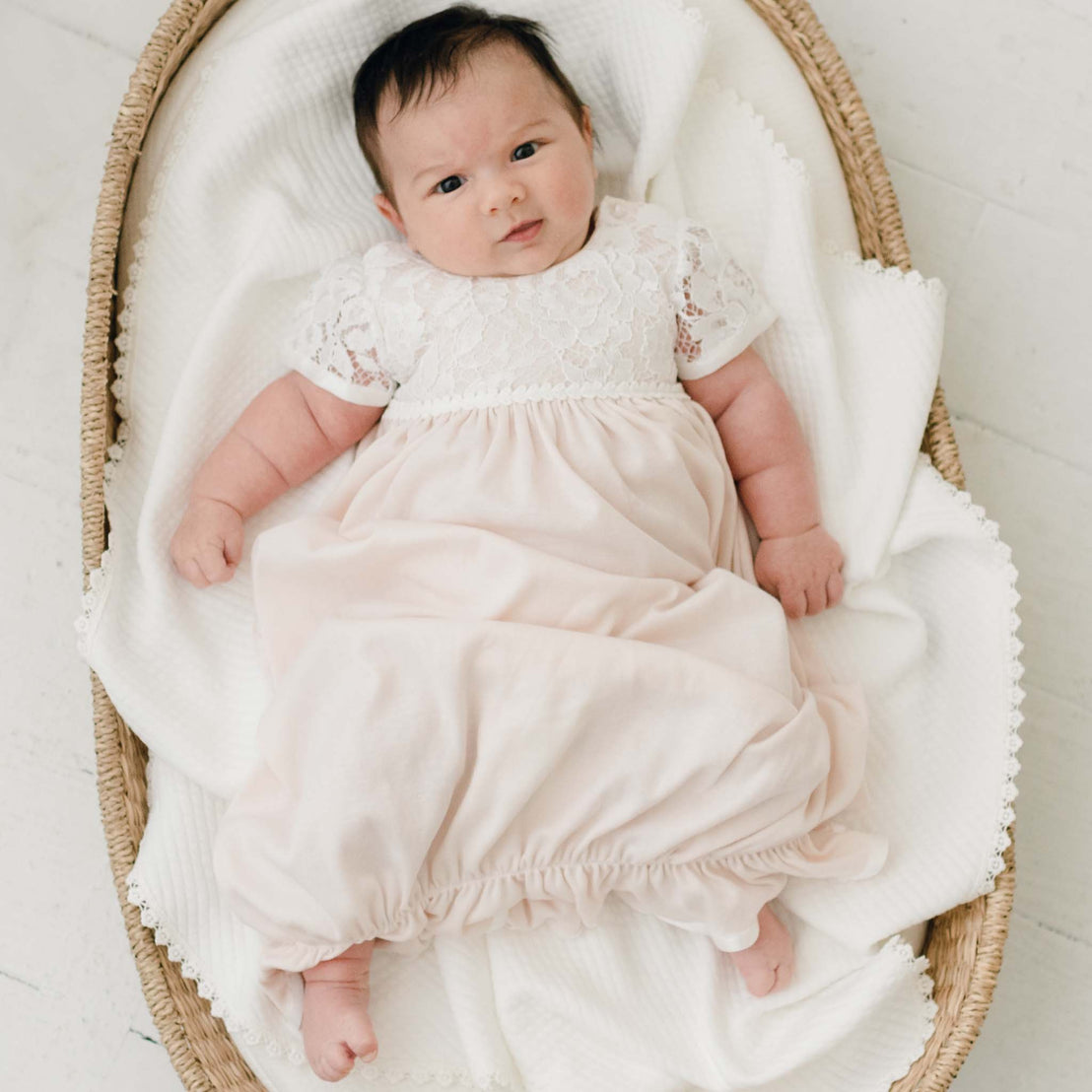 A baby wearing the Rose Layette & Bonnet and lying in a woven basket, looking directly at the camera with a subtle smile.