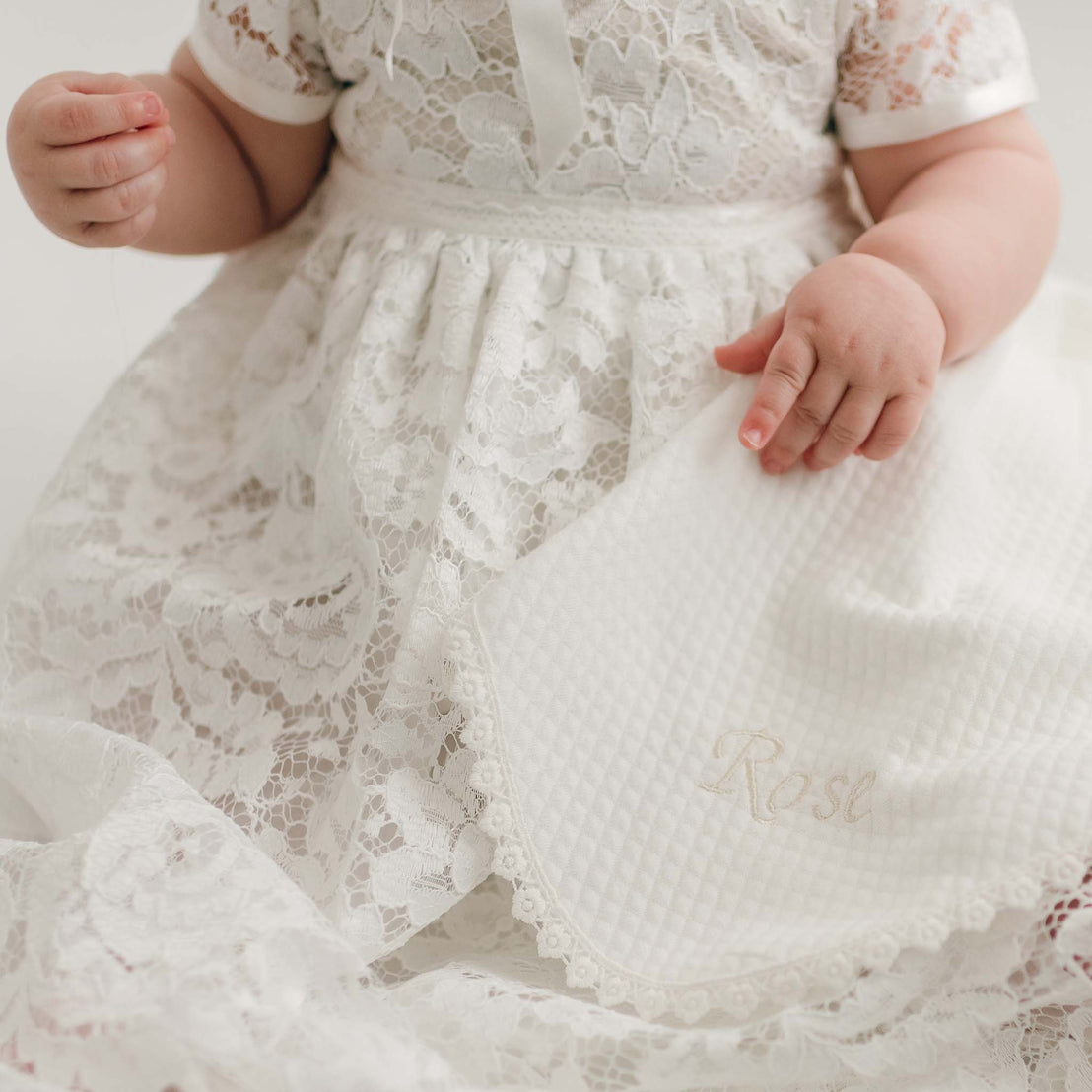 A baby in the Rose Christening Gown holds a Rose Personalized Blanket embroidered with the name "Rose" in a softly lit setting.