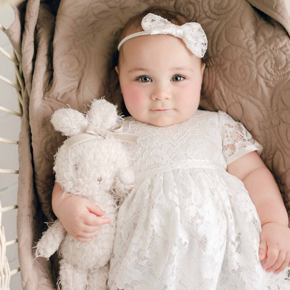 A baby wearing the Rose Romper Dress and matching Rose Bow Headband lies in a wicker bassinet with the Rose Bunny plush toy at her side, looking directly at the camera.