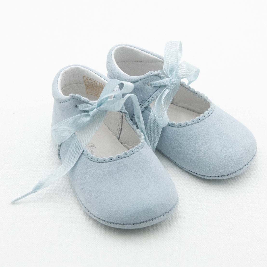 A pair of Emily Suede Tie Mary Janes in light blue, from Baby Beau & Belle, feature delicate ribbon ties and scalloped edging around the opening. The handmade baby shoes are displayed against a plain white background, showcasing their gentle design and craftsmanship.