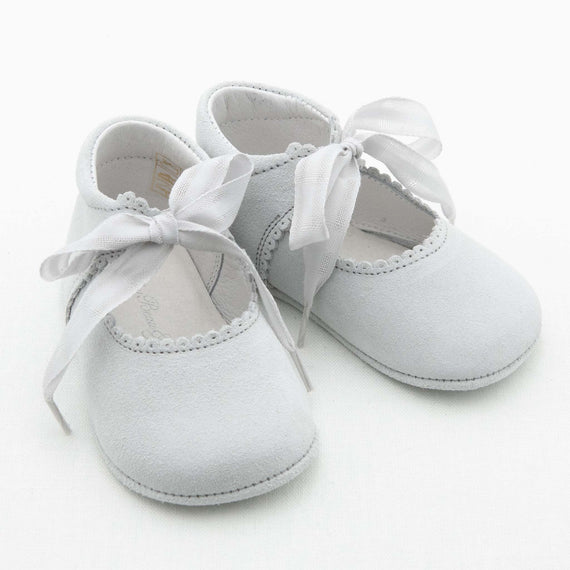 A pair of small, grey Dove Grey Suede Tie Mary Janes baby shoes with soft suede material and ribbon ties. The shoes have a scalloped edge around the opening and appear delicate and comfortable, ideal for a baby. The background is plain white.