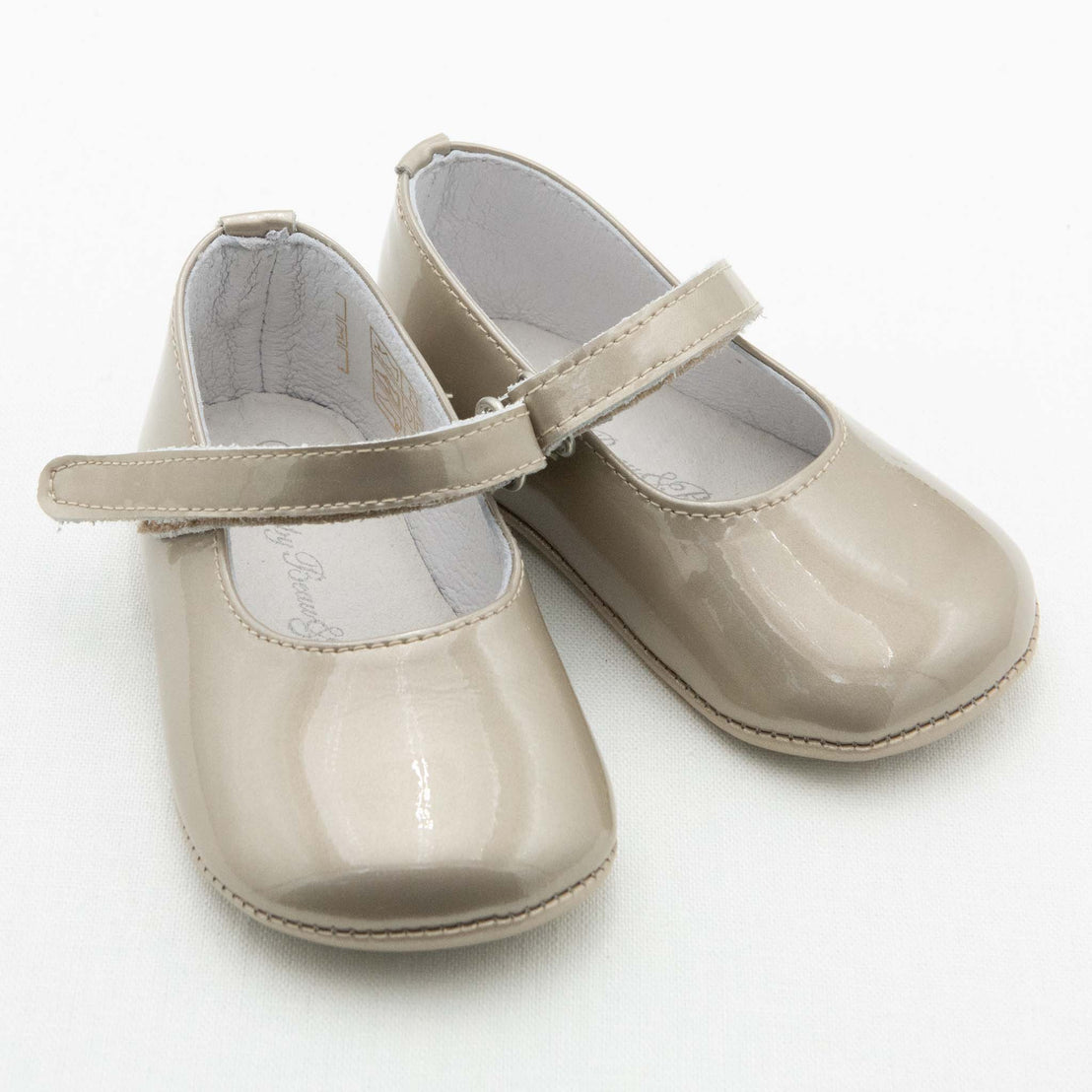 A pair of small Gold Patent Leather Mary Janes. The shoes are made with gold patent leather with a soft velcro strap. The coordinating champagne chiffon bow and feather are removed from the strap. The background is white.