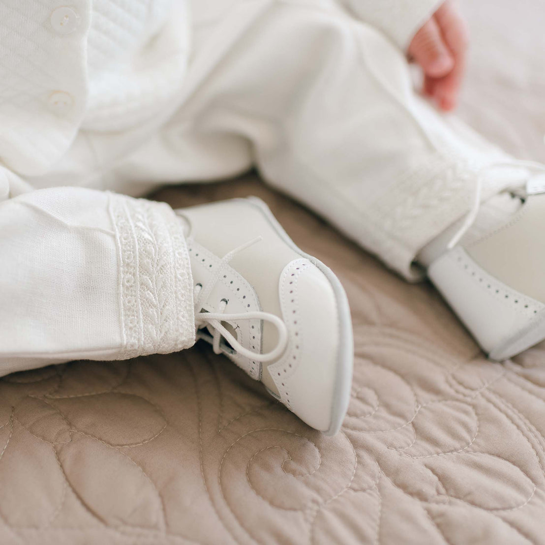 Close-up of a baby's legs and feet wearing the Ivory Two Tone Wingtip Shoes and the Oliver Ivory Suit Pants. The baby is sitting on a beige quilted fabric surface.