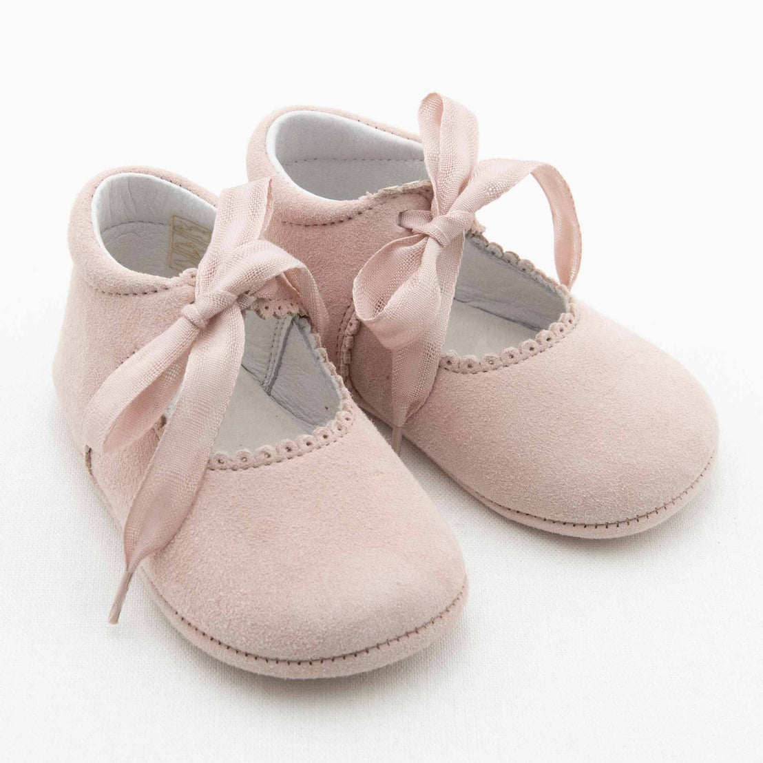 A pair of small, soft pink Eloise Suede Tie Mary Janes with a suede texture and fabric ribbons tied in bows on top. These handmade shoes have scalloped edges around the opening and appear to be designed for infants. Sitting on a white background, they exude the charm of Baby Beau & Belle's signature style.