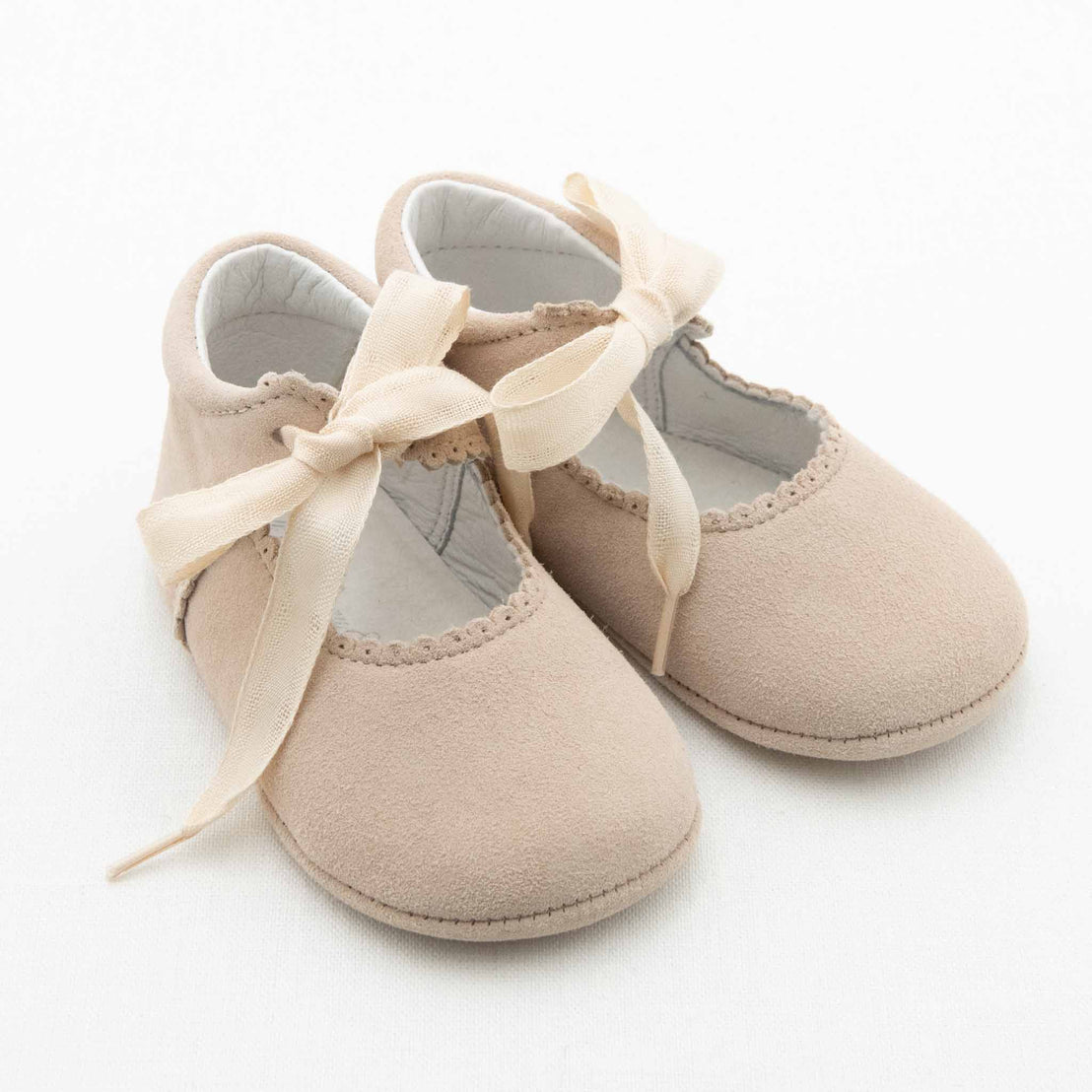 A pair of tan Baby Mary Janes made of soft suede leather with cream ribbon ties. The handmade baby shoes feature a delicate scalloped edge along the opening and appear to be placed on a white surface.