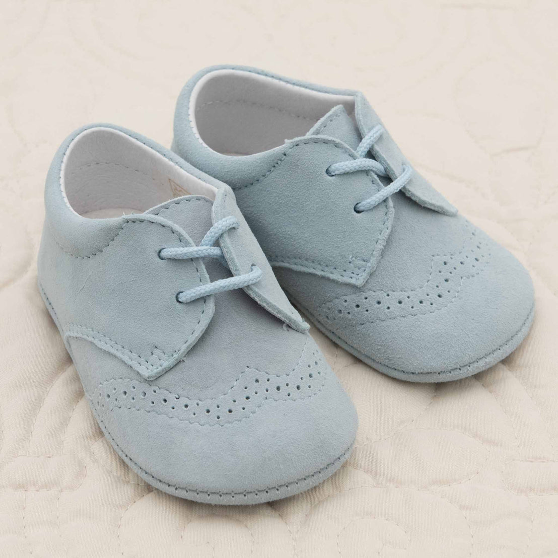 A pair of small, light blue, handmade Asher Suede Shoes made of soft suede featuring lace-up fronts and decorative perforations are placed on a light, quilted surface.