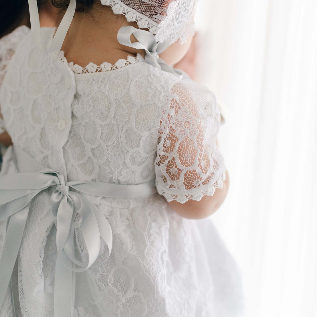 A close-up view of a young girl dressed in the Olivia Dress, perfect for baptism, with short sleeves and a light blue ribbon at the back, standing near a window.