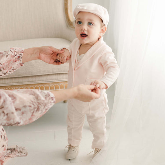 Baby boy standing in blush suit, newsboy cap and suede shoes holding hands