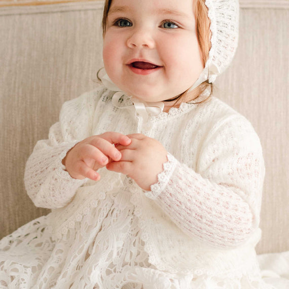 Baby girl wearing the Lola Knit Sweater over the matching Lola Christening Gown. The sweater is made from soft, breathable light ivory knit, featuring button closures and ivory trim.