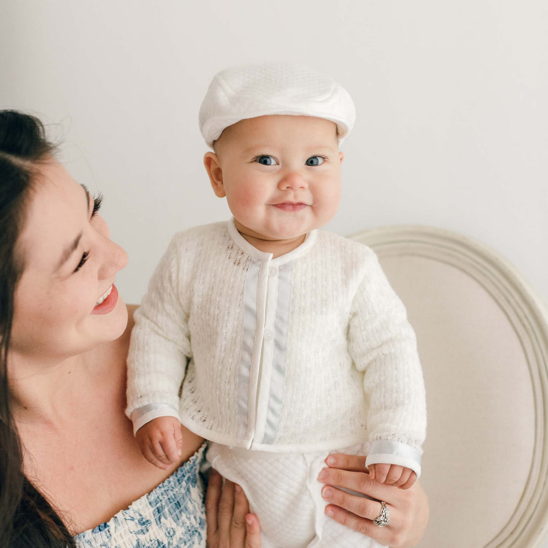 Smiling baby boy standing on a chair with his mother nearby. He is wearing the Owen 3-Piece Suit, including the sweater, pants, and onesie