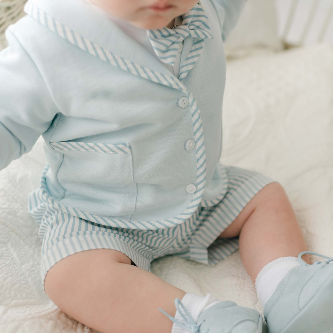 A baby dressed in a Theodore Shorts Suit, made from soft French Terry cotton, sits on a white quilted blanket. The pale blue outfit is accented with white and blue striped details on the collar, bow tie, and shorts. The ensemble is completed with matching blue shoes and white socks. The baby's face is partially out of the frame.