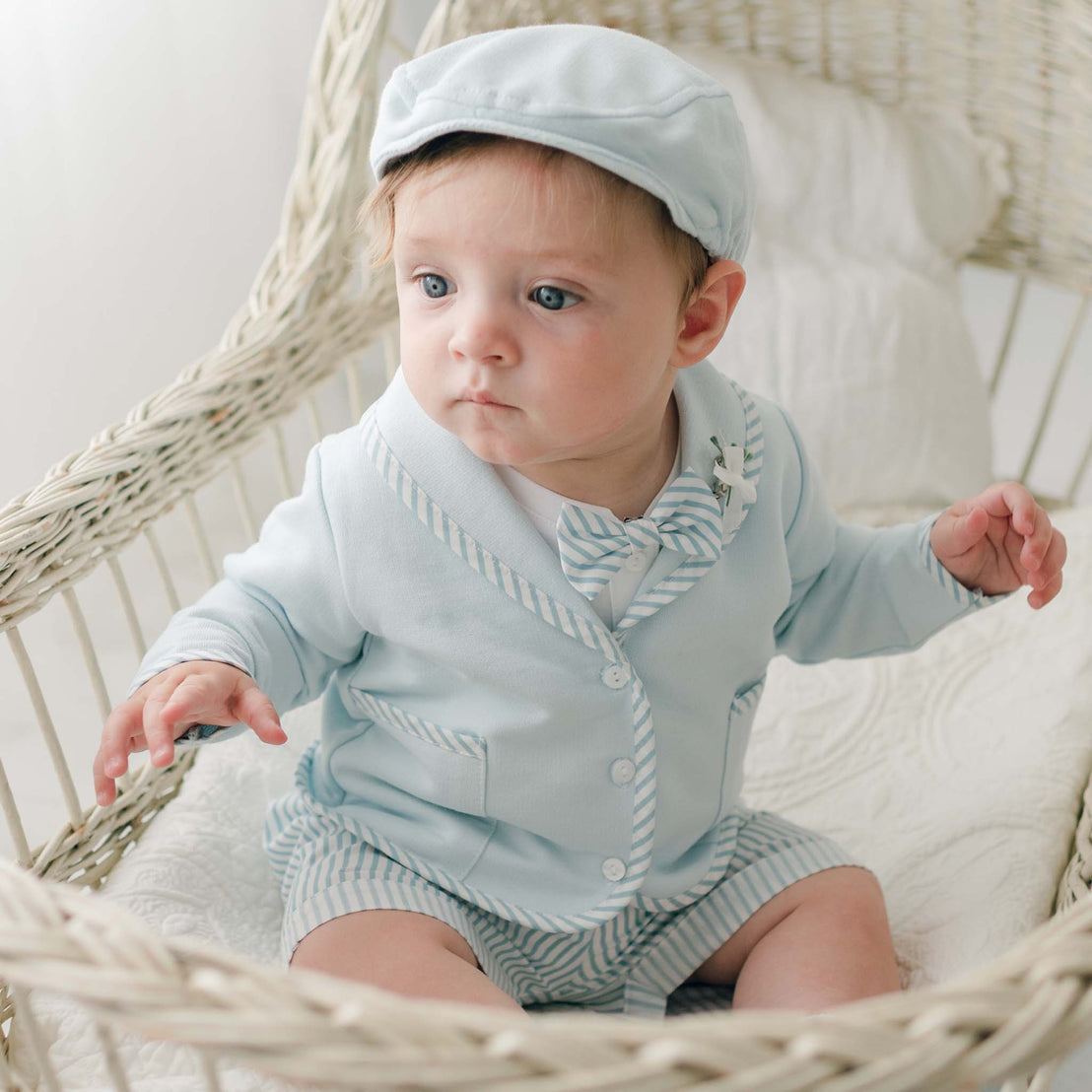 A baby with light hair, dressed in a Theodore Shorts Suit made of French Terry cotton, sits in a white wicker chair. The handmade suit features a light blue jacket with striped trim, matching shorts, and a bow tie. The baby looks off to the side with a curious expression.