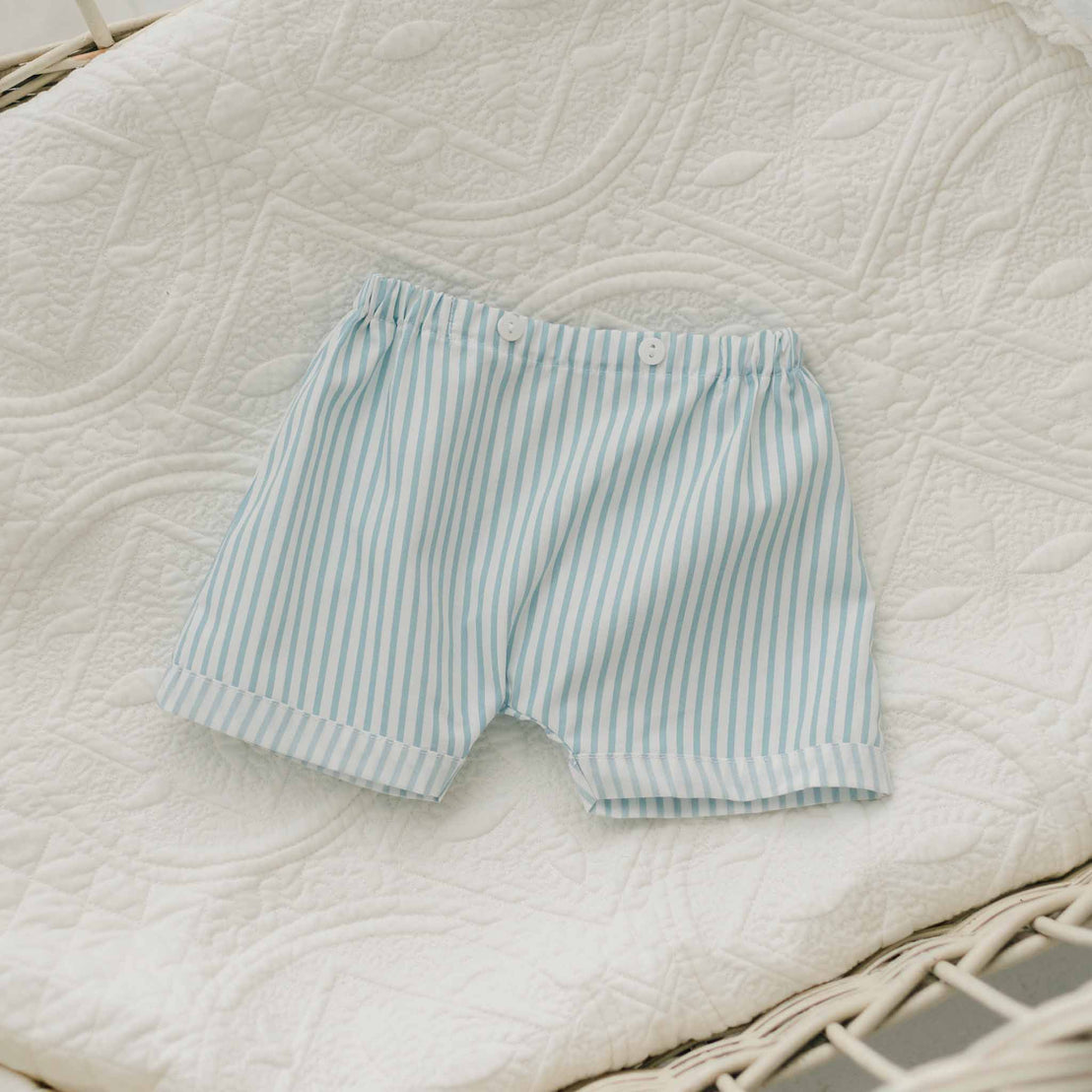 A Theodore Shorts Suit, designed for baby boys in light blue and white stripes, featuring an elastic waistband and two small front buttons, crafted from soft French Terry cotton and showcased on a quilted white blanket with a decorative pattern.