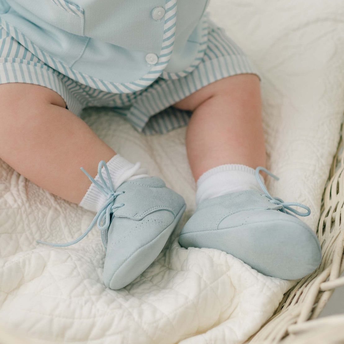 Close-up of a baby's legs and feet. The baby is wearing a light blue, short-sleeved outfit with stripes, white socks, and Theodore Suede Shoes in light blue suede. The baby is sitting on a quilted white blanket inside a wicker basket.