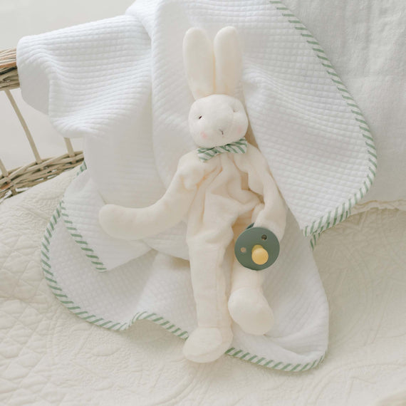 Theodore Bunny Buddy, a plush toy crafted from soft velour and featuring a green pacifier attached to its hand, rests on a white quilted blanket with green trim. The scene is set in a cozy and light-colored environment, perhaps in a crib or cradle—perfect as an ideal baby gift.