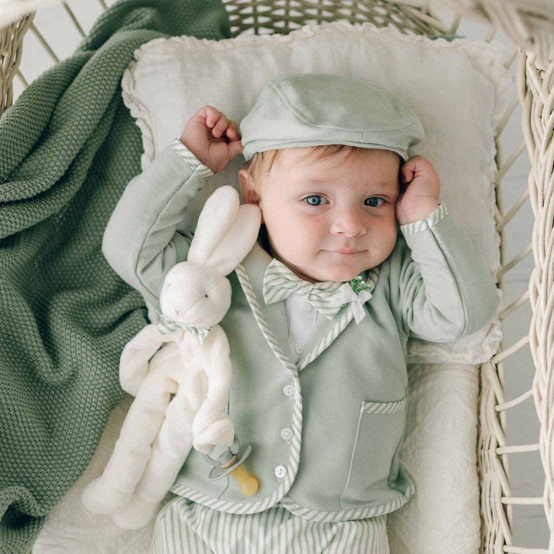 A baby dressed in a light green cap, jacket, and striped pants lies on a white pillow in a wicker basket. The baby is holding the Theodore Bunny Buddy | Pacifier Holder. A soft velour green blanket is draped next to the baby.