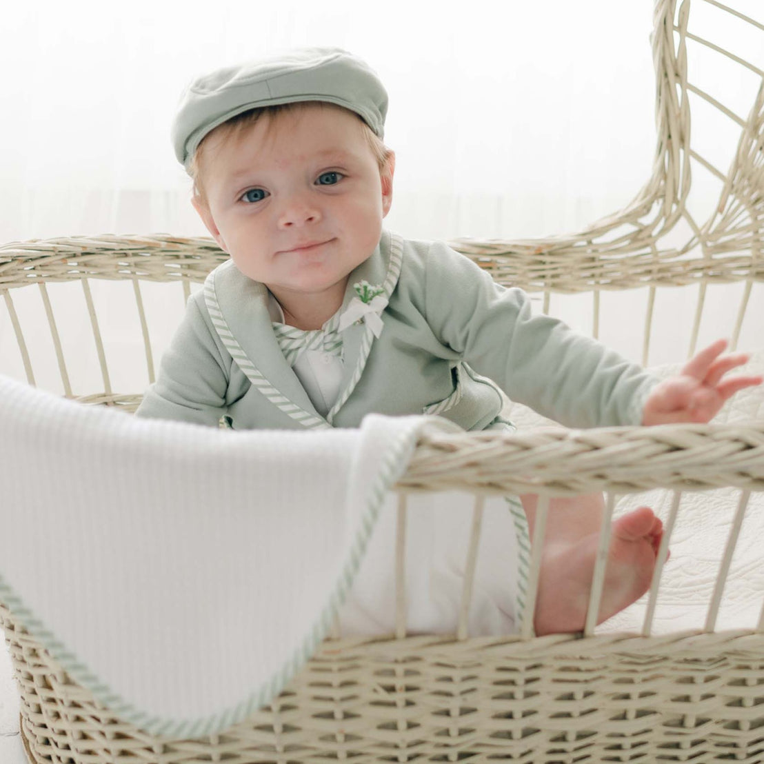 A baby wearing a mint green Theodore Shorts Suit and matching cap sits in a woven bassinet. The French Terry cotton suit complements the white blanket with a green trim draped over the side. The baby has a curious expression and one hand lifted against the softly lit background.