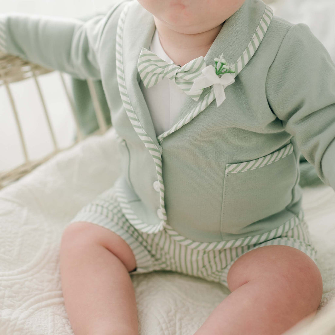 A baby sits in a light-colored wicker crib, wearing a light green Theodore Shorts Suit featuring a blazer with striped trim and matching shorts. The soft French Terry cotton blazer has a small decorative floral pin on the lapel, and the baby is holding onto the side of the crib.