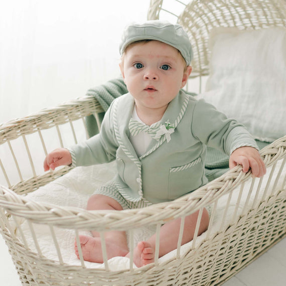 A baby dressed in an elegant mint green Theodore Shorts Suit, complete with a cap, sits inside a white wicker bassinet lined with soft blankets and green fabric. The baby has a curious expression and light eyes, with soft natural light illuminating the scene.